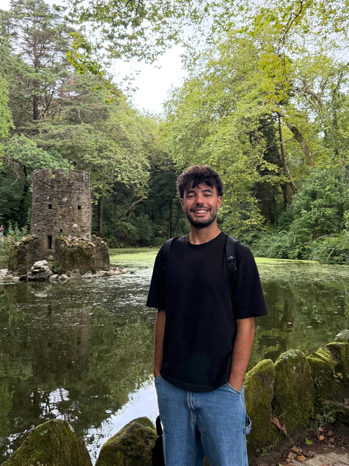 Raúl smiling by a pond in a forest, an old stone tower behind him